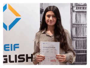 Estudiante joven con pelo largo negro y camisa de rayas sonríe satisfecha en nuestra academia de inglés de Madrid, enseñando su certificado de aprobado del Aptis C1.