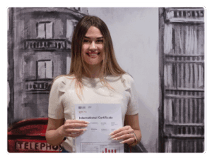 Estudiante joven con pelo largo y camiseta blanca sonríe satisfecha en nuestra academia de inglés de Madrid, enseñando su certificado de aprobado del Aptis B1-B2.