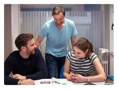 Profesor nativo en aula moderna ayudando a dos estudiantes del curso de inglés general con el libro Oxford English File, uno de los manuales más prestigiosos